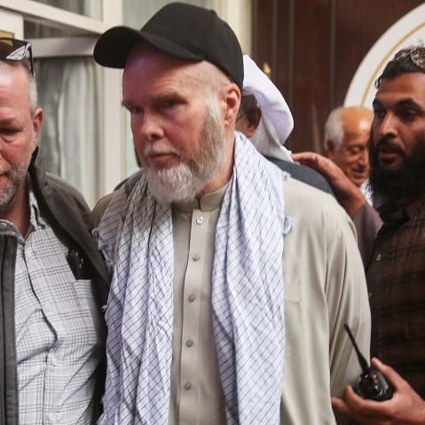 Dennis Coyle looks away, wearing a traditional Afghani clothing with a baseball cap as her is escorted by two men.
