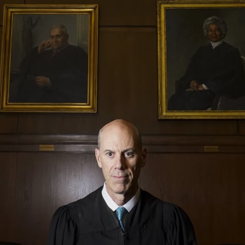 A judge in his robes stands beneath eight gold-framed portraits of other judges