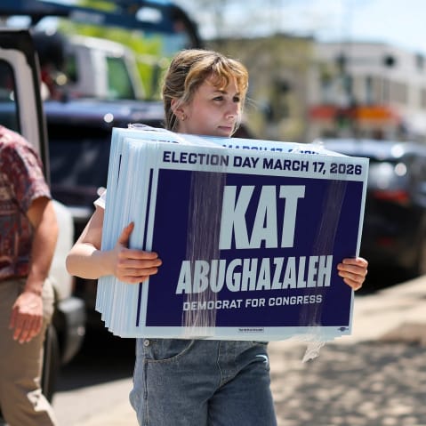 Kat Abughazaleh carries her signs on the sidewalk while a man walks towards a van in the background.