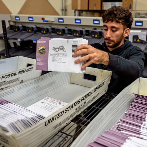 A person grabs a stack of mail-in ballots from a United States Postal Service tray of ballots.