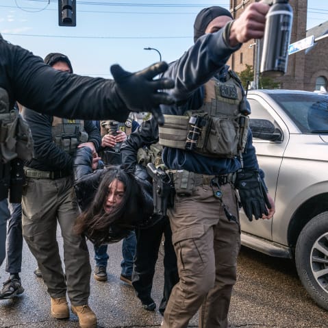 Federal agents hold a person by their arms and legs in the street.