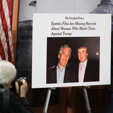 There are two photographers on the left bottom of the frame taking pictures of a poster that shows a photo of Donlad Trump and Jeffrey Epstein together.