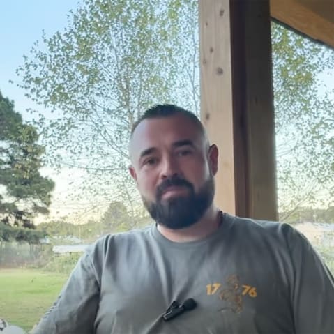 Aaron Spencer sits at a porch as he speaks. Turkeys can be seen in the background.