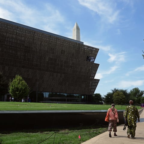 Two people walk by the National Museum of African American History and Culture on a sunny day.