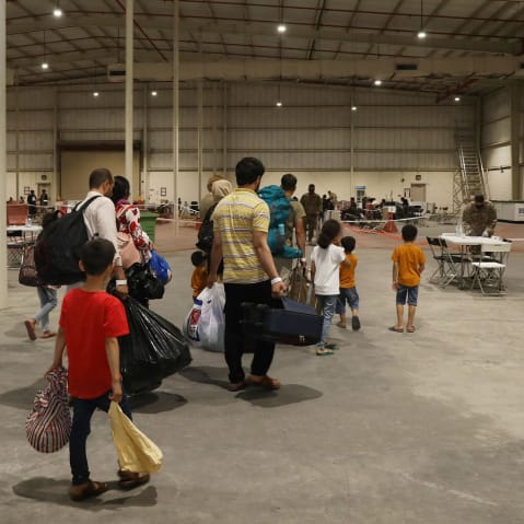 People hold their belongings while walking through a big warehouse-like building with tables and chairs monitored by people in army uniforms.