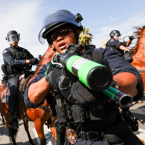 A police officer holds a weapon to the camera as protesters face off with police outside of a federal building in downtown Los Angeles for an anti-Trump "No Kings Day" demonstration.