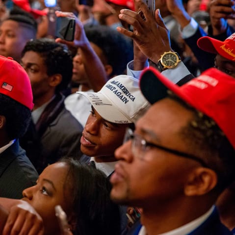 Audience members, most of them Black, wear MAGA hats and hold up their phones to take photos.