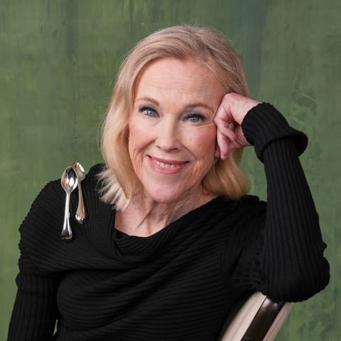 Catherine O'Hara in all black with two spoons on her shoulder. She sits in front of a painted green backdrop.