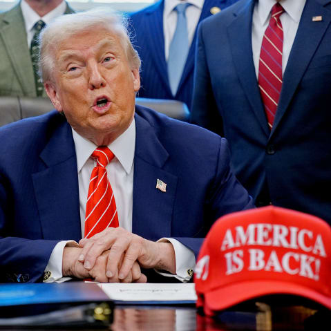 President Donald Trump, center, surrounded by members of the House and Senate in the Oval Office. Out of focus on the desk is a red hat that says "AMERICA IS BACK!"