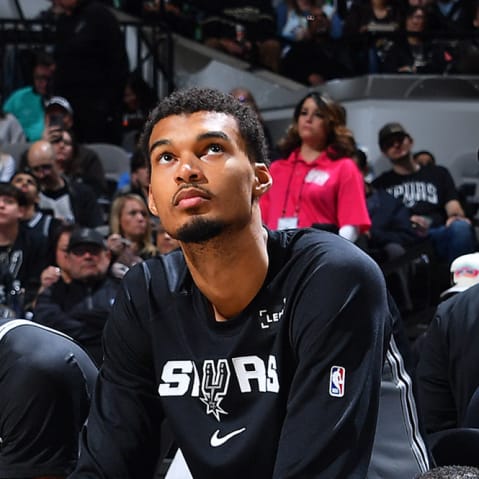 Victor Wembanyama, center, sits on the sidelines of a basketball court surrounded by teammates.