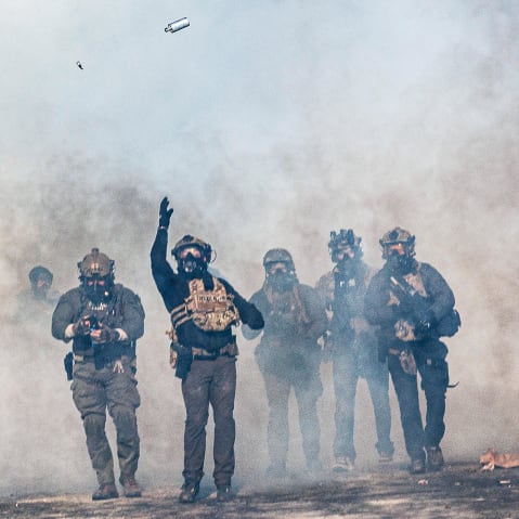 One federal agent, walking through a cloud of tear gas, throws a teargas canister which is now suspended in midair. Other federal agents walk alongside.