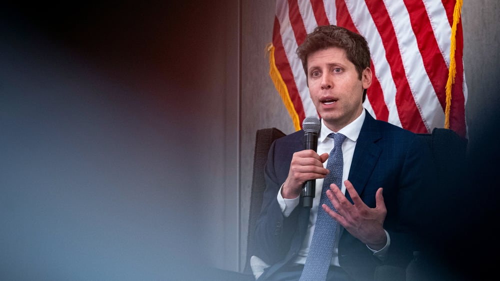 Sam Altman holds a microphone. An American flag is partially visible behind him.
