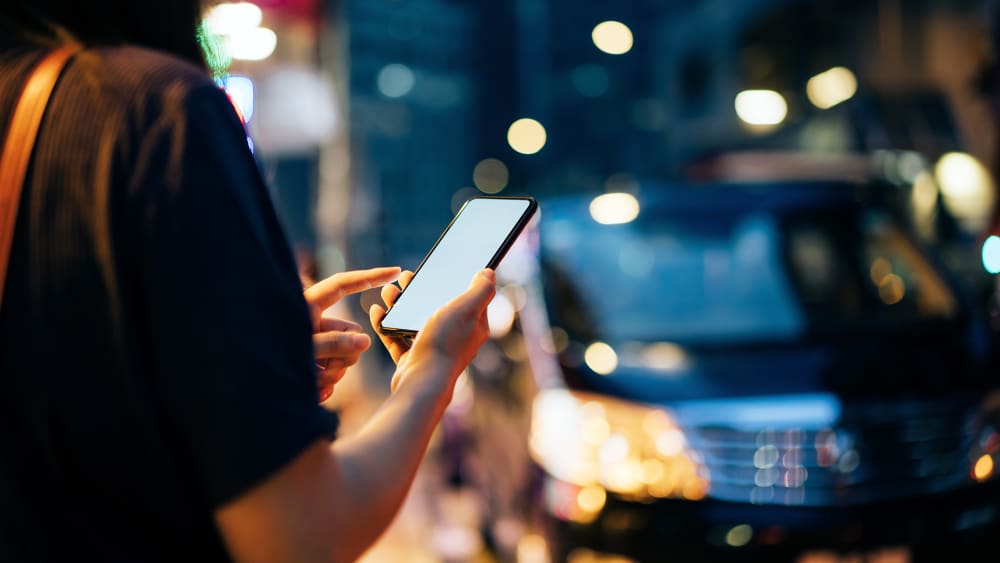 Close up of young woman using mobile app device on smartphone to hail a taxi ride on city street after work in the evening.