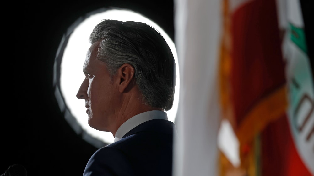 Gavin Newsom backlit, next to a California state flag.