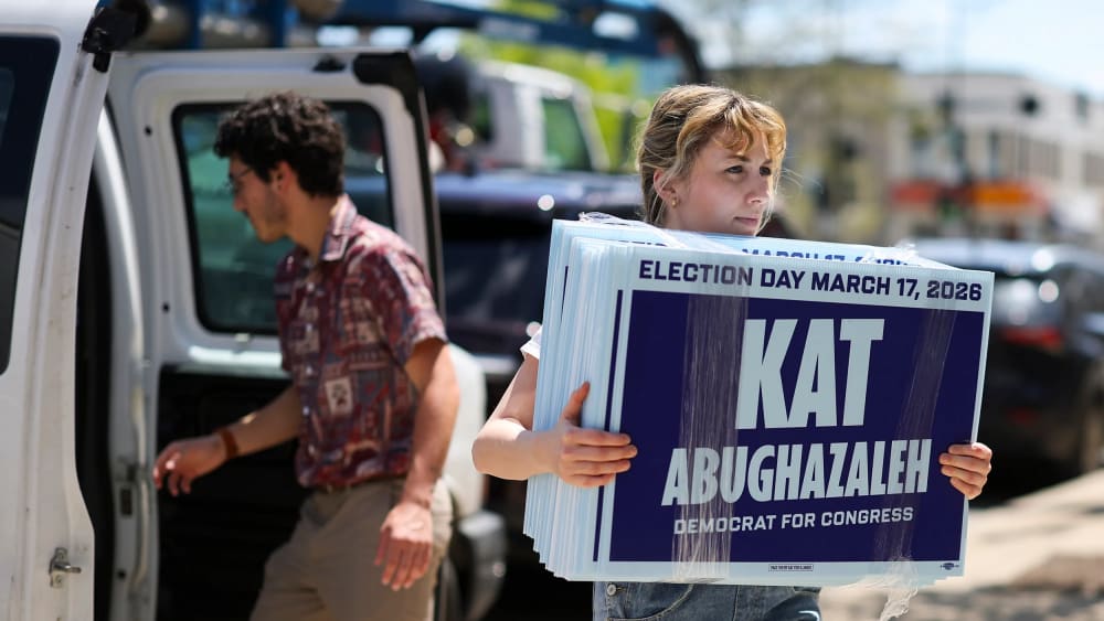Kat Abughazaleh carries her signs on the sidewalk while a man walks towards a van in the background.