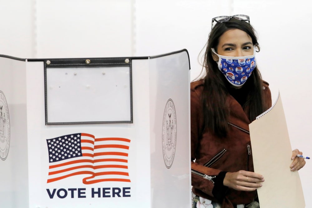 Image: Alexandria Ocasio-Cortez stands next to her voting booth holding her filled ballot.