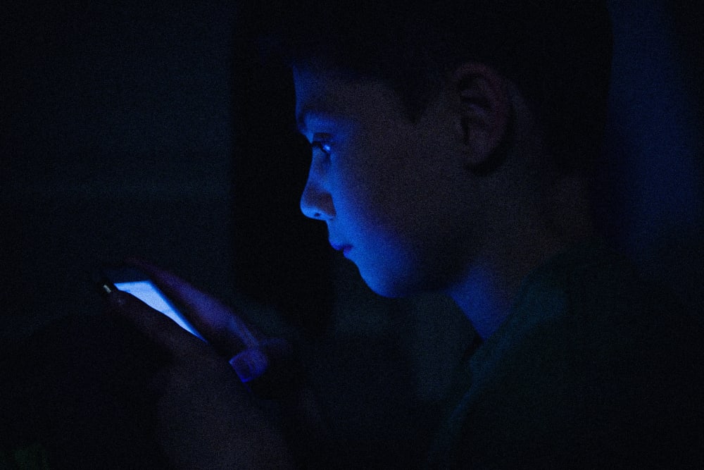 IMage: a teenage boy on his phone, face illuminated in the dark