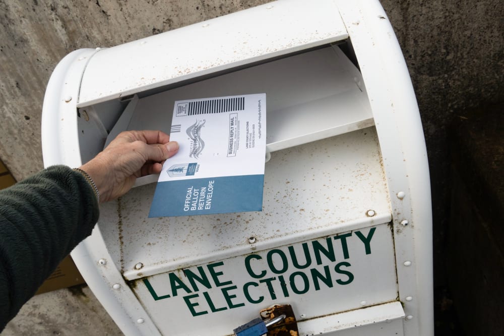 A hand hovers a ballot over a ballot drop box