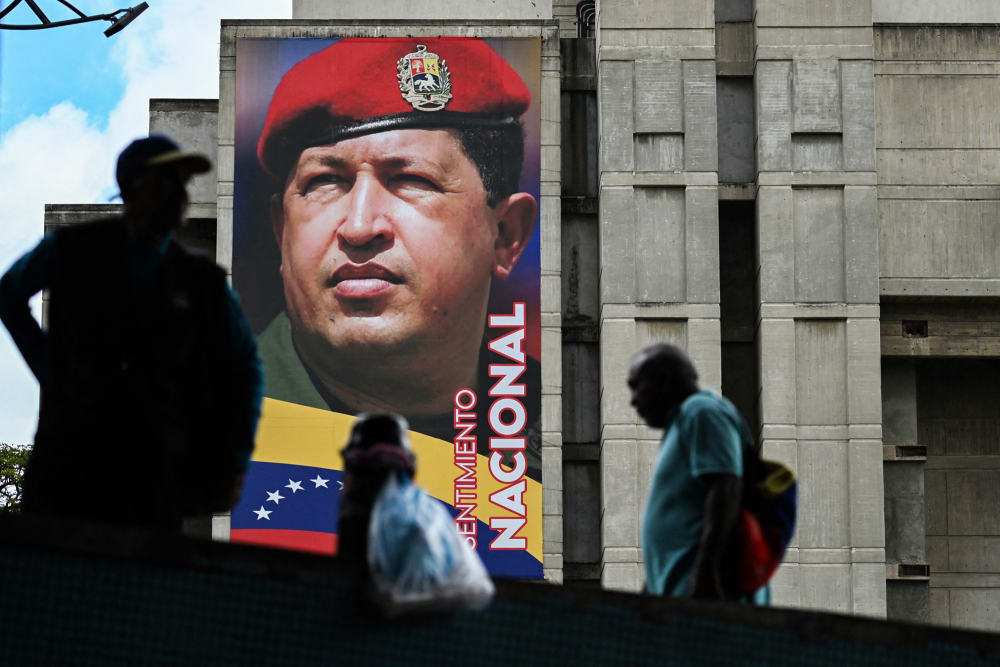 People walk past a mural of Hugo Chavez on the side of a building outside