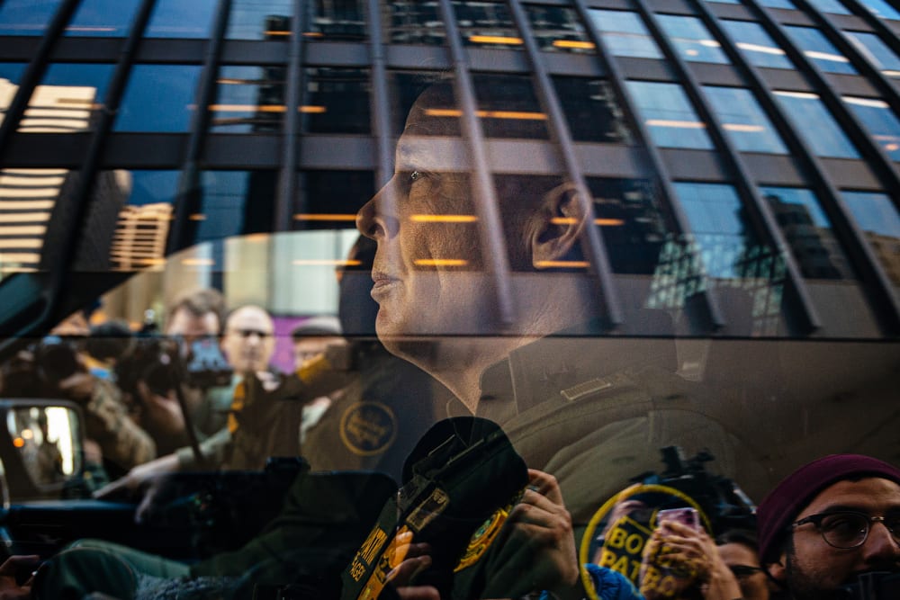 U.S. Border Patrol commander Gregory Bovino sits in the driver's seat surrounded by media and protesters.