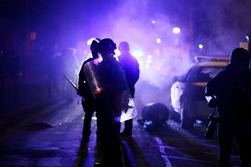 In this Nov. 25, 2014 file photo, police officers watch protesters as smoke fills the streets in Ferguson, Mo. after a grand jury's decision in the fatal shooting of Michael Brown. (Photo by Charlie Riedel/AP)