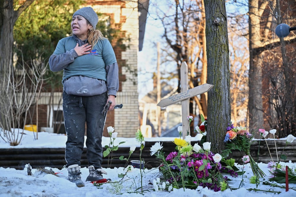A woman in distress with her hand on her heart stands next to a wooden cross surrounded by flowers.