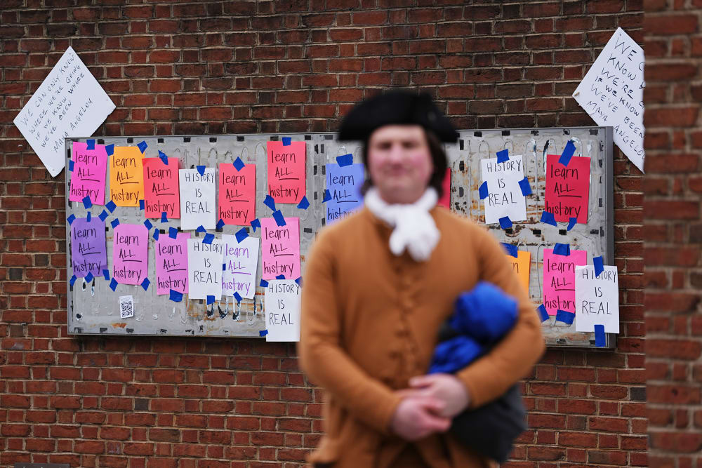 A person dressed in a late 18th- or early 19th- century costume stands blurred in the foreground. In focus in the background, multicolored paper signs that read "learn ALL history" and "HISTORY IS REAL" are taped to an empty panel on a brick wall.