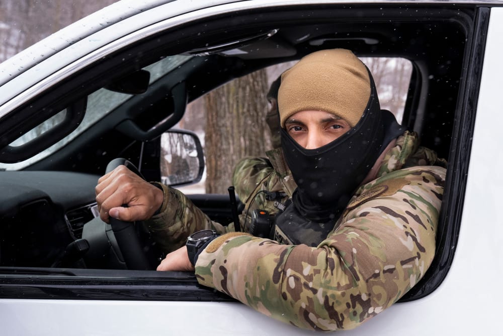 A masked agent driving a white car looks out the window of the car.