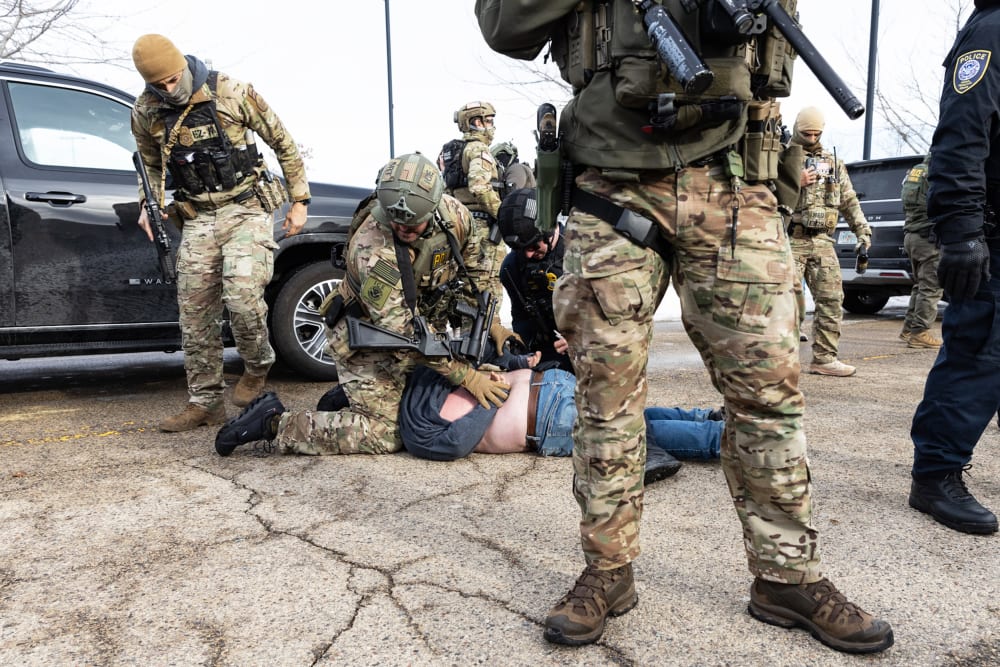 An ICE agent sits on top of a person, who is face down on the ground, and pins the person&rsquo;s hands behind their back. Other agents stand nearby.