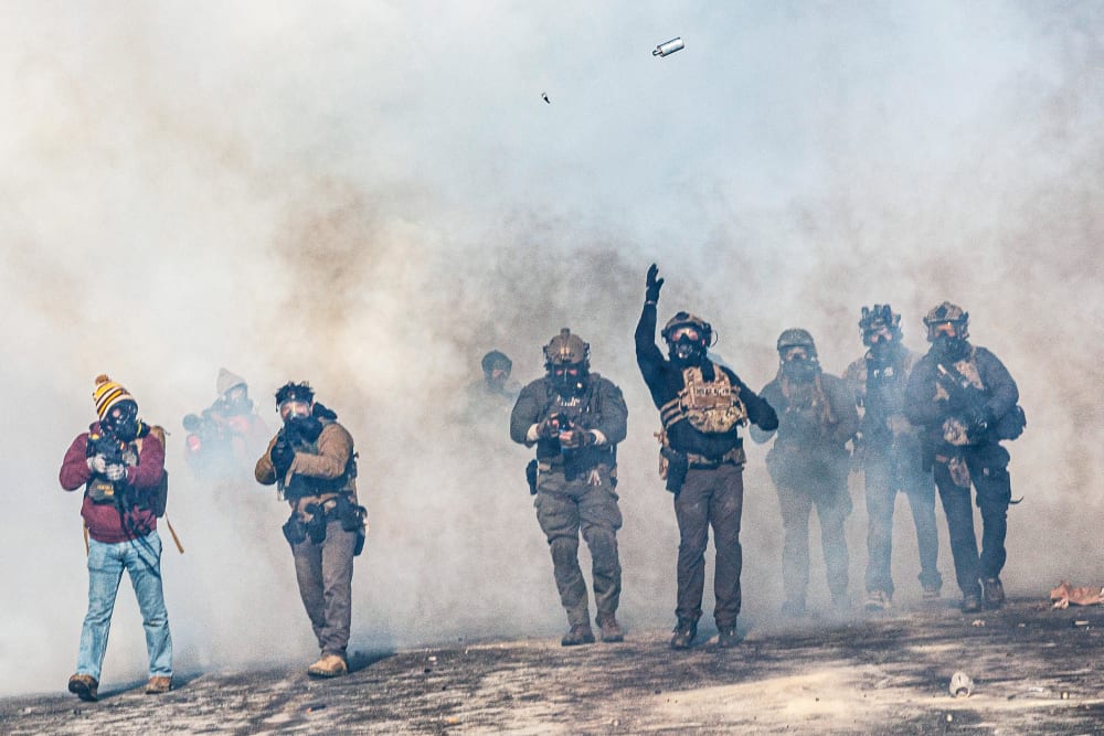 One federal agent, walking through a cloud of tear gas, throws a teargas canister which is now suspended in midair. Other federal agents walk alongside.