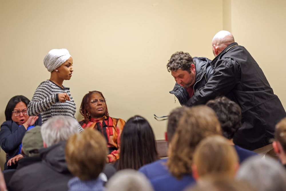 Ilhan Omar, left, raises her fist as if to throw a punch while security tackles a person from behind.