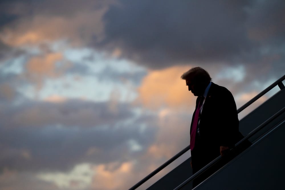 Donald Trump, silhouetted, descends an outdoor staircase. A sky full of colorful clouds is behind him.