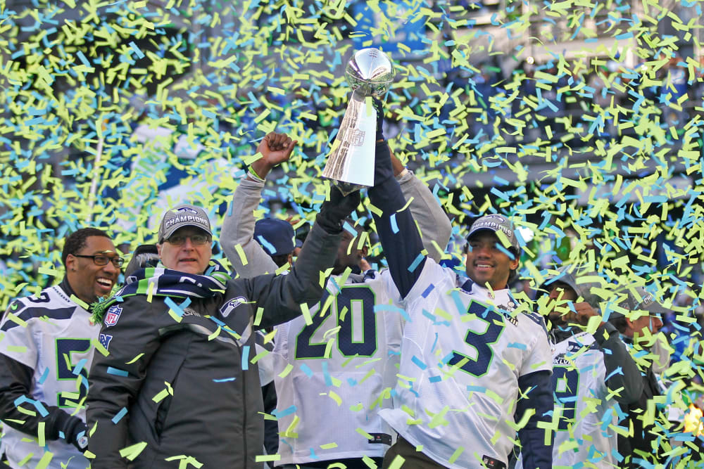 Paul Allen, left, and Russell Wilson hoist the Lombardi trophy in the air as blue and green confetti falls around them.