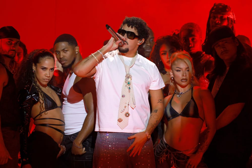 Bad Bunny, surrounded by performers, holds a microphone against a red background.