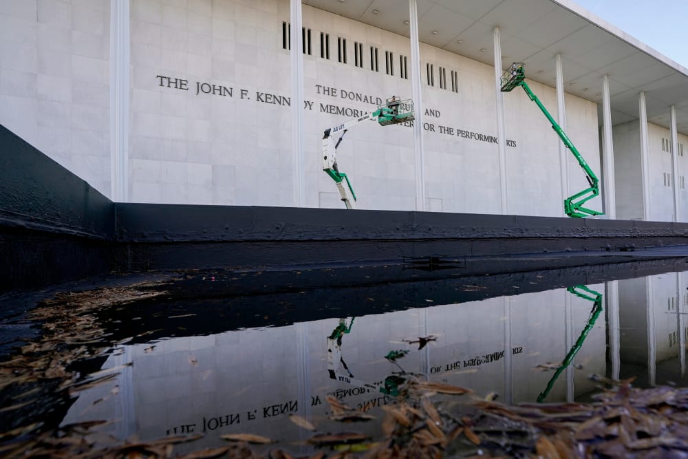 Workers in green construction lifts work on the facade of the Kennedy Center, which is reflected in a puddle.