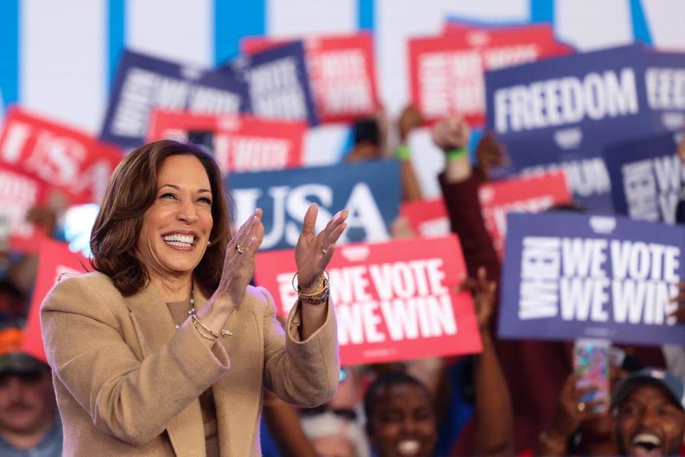 Kamala Harris claps; people behind her hold up signs in support.