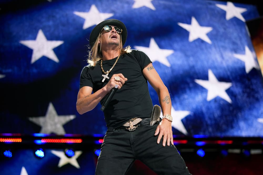 Kid Rock holds a microphone on stage. The background is an American flag.