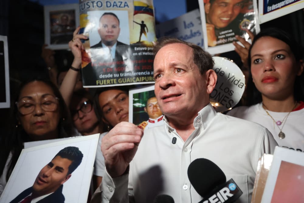 Juan Pablo Guanipa stands in front of a microphone with a crowd of people behind him holding signs for the release of other Venezuelan prisoners.