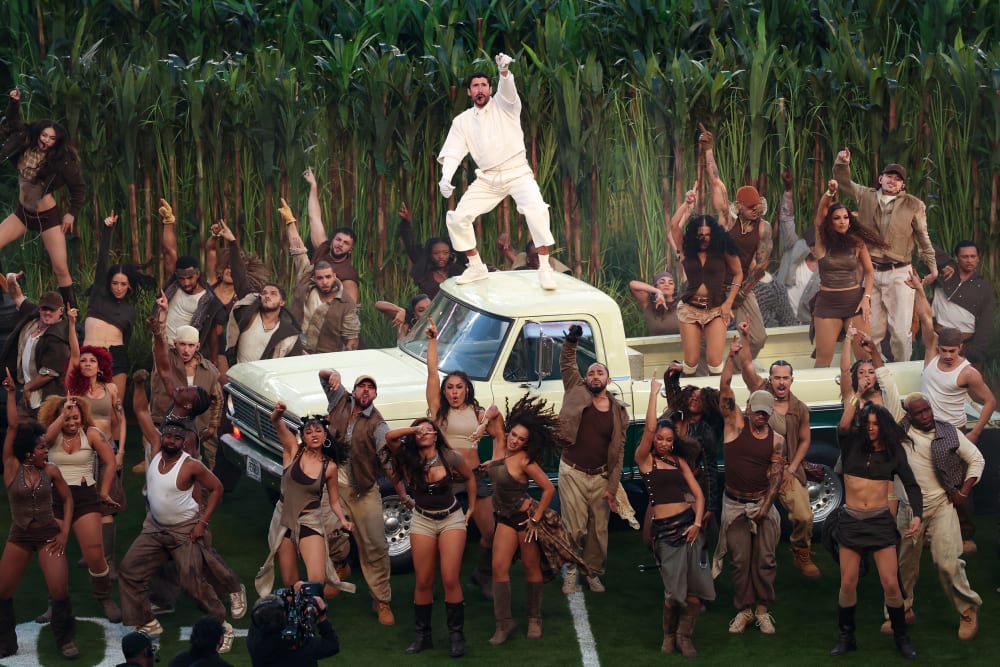 Bad Bunny stands on the roof of a light yellow truck. People dance in the truck bed and on the football field below.