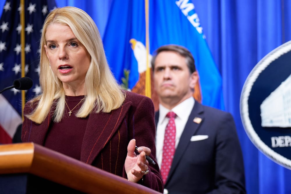 Pam Bondi stands behind a podium and microphone; Todd Blanche stands behind her, out of focus.