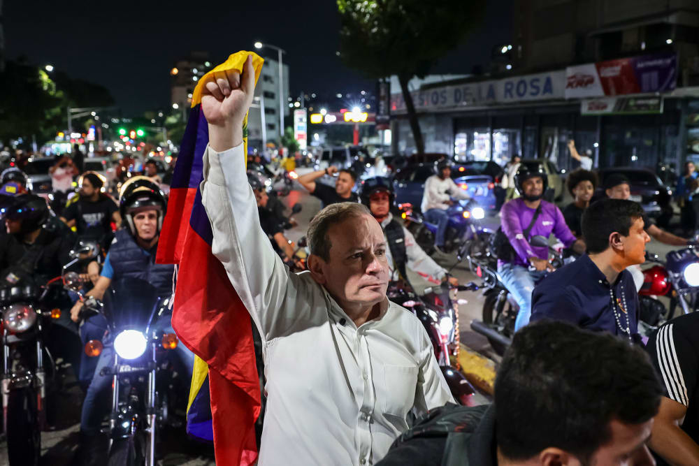 Juan Pablo Guanipa, center, stands in the street, holding a Venezuelan flag up with one hand. He is surrounded by people on motorcycles.