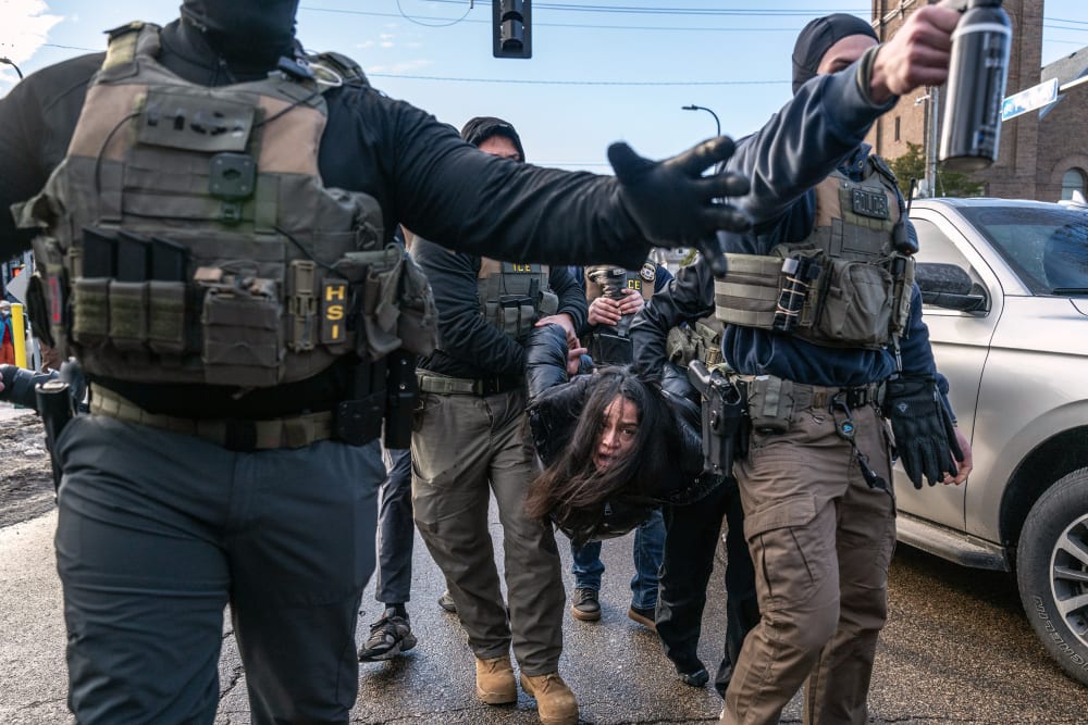 Federal agents hold a person by their arms and legs in the street.