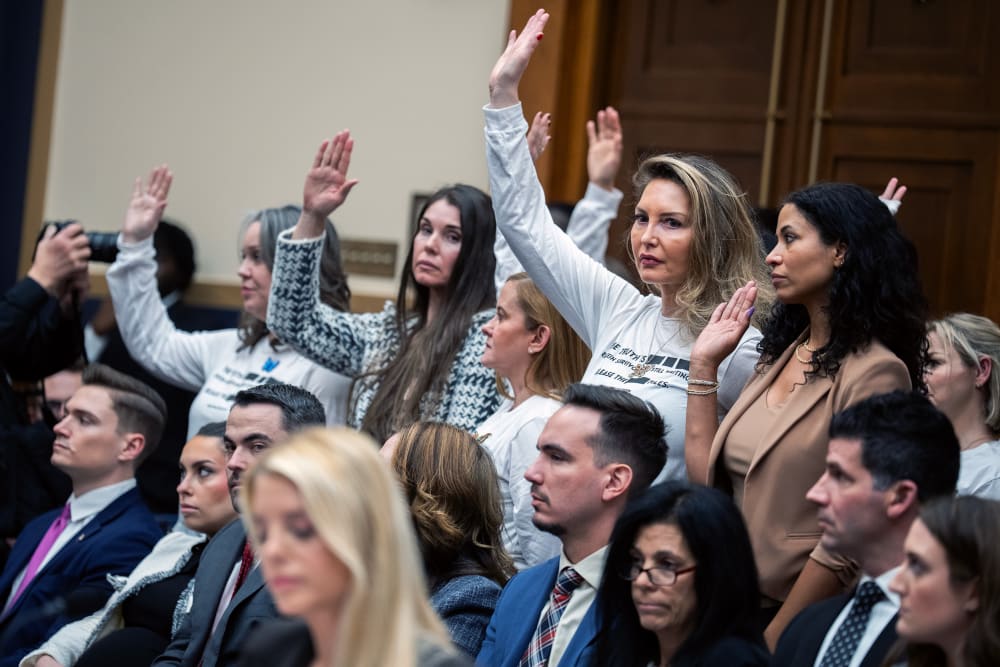 People in focus in the background raise their hands; Pam Bondi out of focus in the foreground looks down.