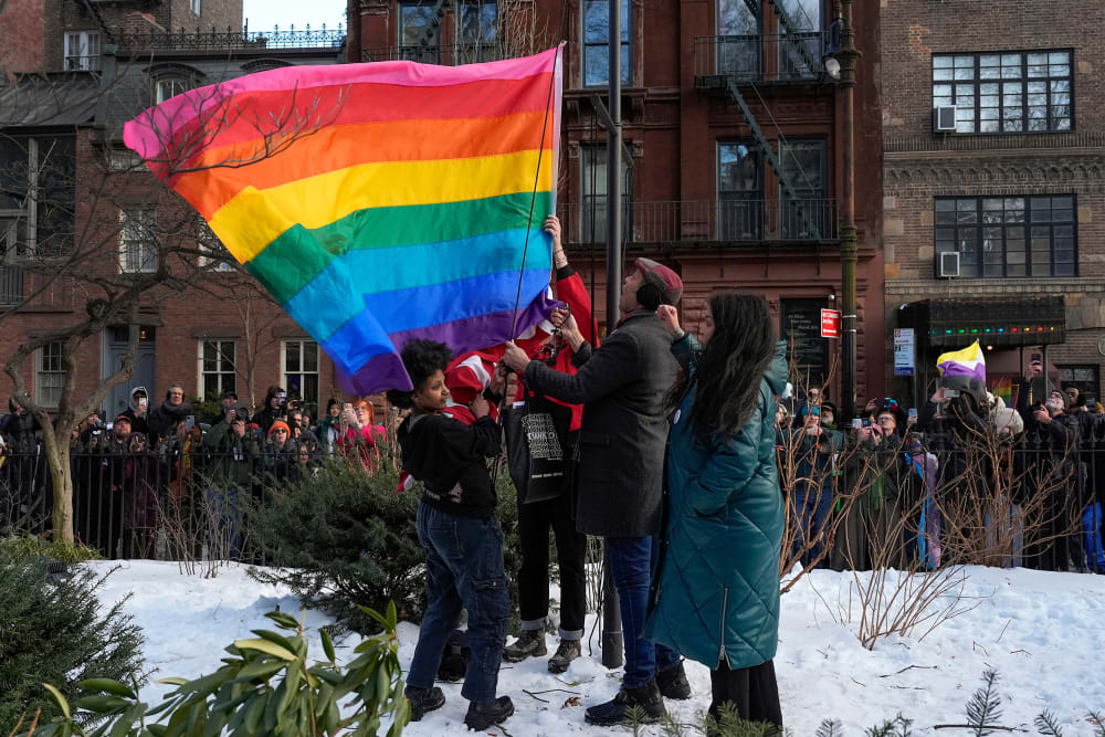 People raise a rainbow flag on a pole in a park. A crowd is gathered behind, watching and filming.