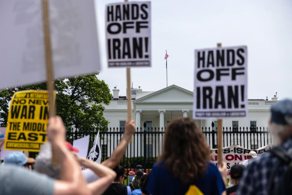 Out of focus in the foreground, people hold picket signs that read "Hands Off Iran!" In focus in the background is the White House.