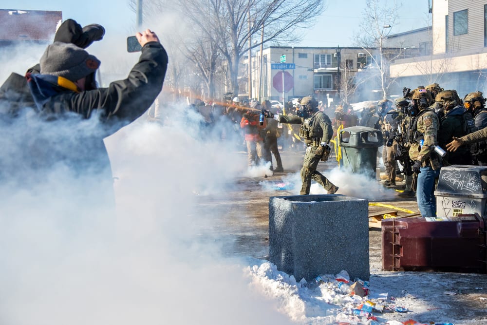 A person on the left films while a federal agent, center, sprays an orange substance ahead. On the right, other agents standby.
