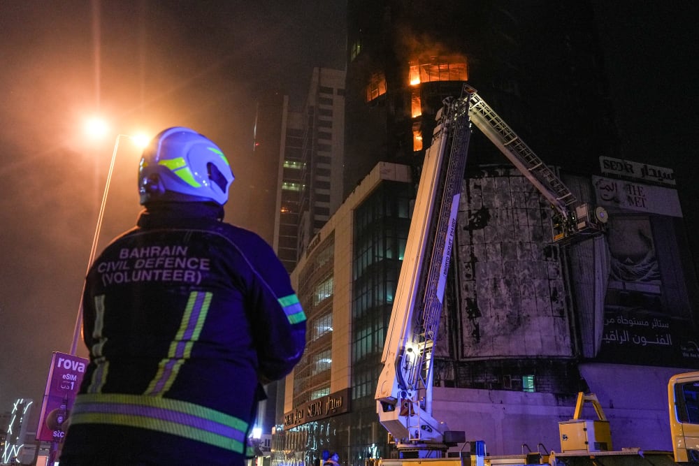 A person in a hard helmet with a jacket that says "Bahrain Civil Defence (Volunteer)" looks up at a building which is on fire.