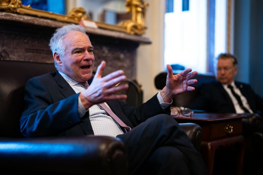 Tim Kaine, in focus on the left, gestures with his hands, while Rand Paul, out of focus on the right, looks at him.