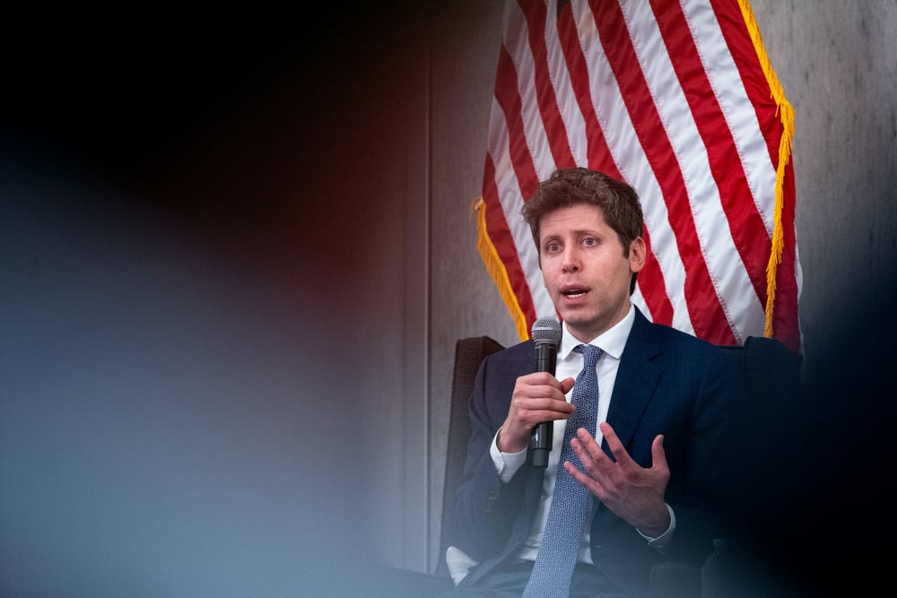 Sam Altman holds a microphone. An American flag is partially visible behind him.