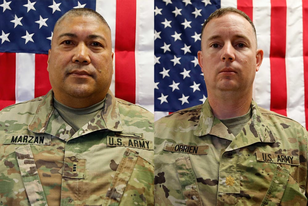 Two people in army uniforms in front of American flags.